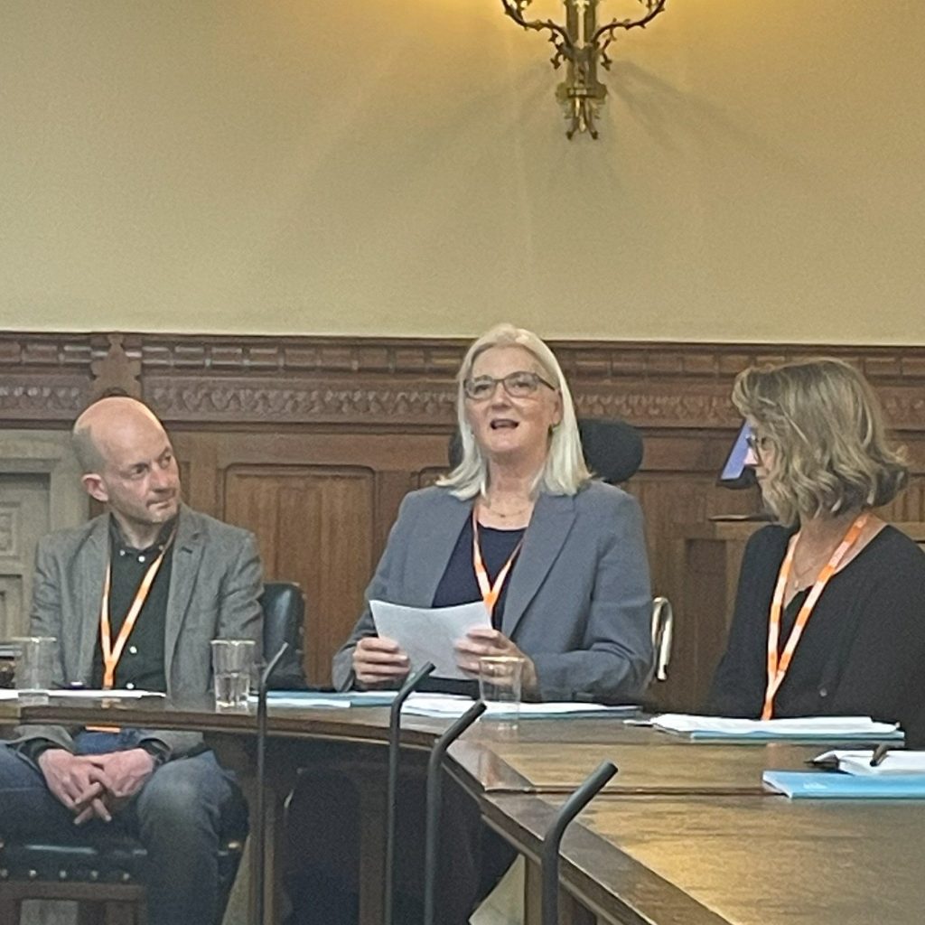 In a parliamentary building, Catherine Hale is sat between 3 colleagues. She is wearing a grey blazer and black top and is presenting to the audience