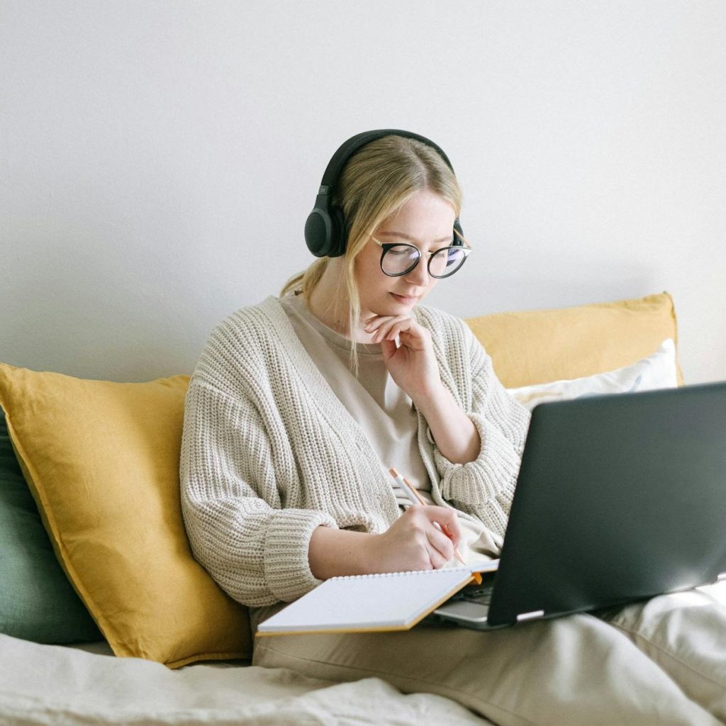 A young white woman in headphones wearing a cost pale jumper sat in bed against some yellow pillows with a laptop and notepad on her lap